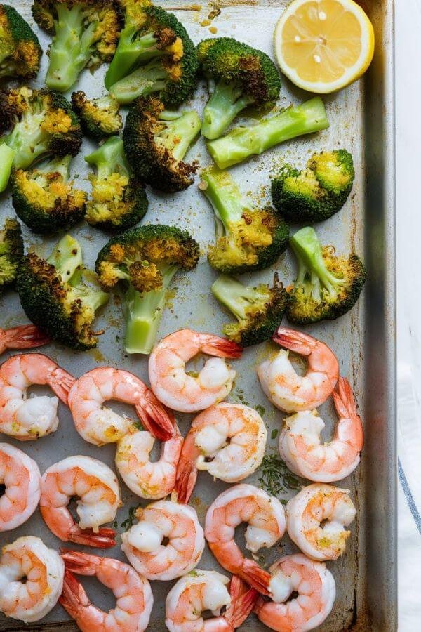 A vibrant sheet pan with bright green roasted broccoli, golden-brown edges, and plump, pink shrimp glistening with olive oil and a squeeze of fresh lemon, all served on a white table with a lemon wedge and a sprinkle of garlic powder on the side.