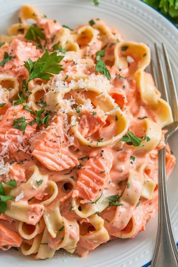 A white plate of creamy salmon pasta with fettuccine, garnished with fresh parsley and some grated Parmesan on top. The salmon flakes are visible, and the creamy garlic sauce glistens under soft, natural lighting. Place a fork resting beside the plate for a cozy, inviting look.