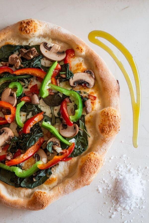 A freshly baked pizza with a golden sourdough crust, topped with vibrant vegetables like spinach, bell peppers, and mushrooms, sitting on a white counter with a drizzle of olive oil and a sprinkle of sea salt nearby.