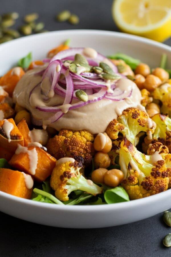 A vibrant, earthy salad in a whitebowl. Roasted sweet potatoes, golden chickpeas, and cauliflower are glistening with light dressing. The red onion adds a pop of color, while a drizzle of tahini dressing pools on top. The background includes scattered pumpkin seeds and a lemon wedge for garnish. Warm tones and natural lighting create a comforting feel.