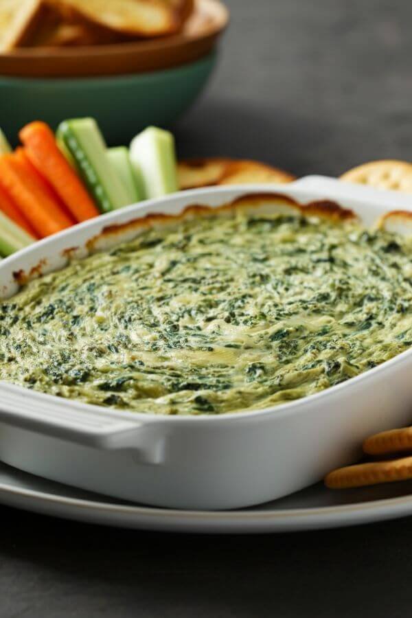 A bubbling baking white dish filled with slight green Spinach and Artichoke Dip, surrounded by a platter of crackers, toasted bread slices, and colorful veggie sticks, with steam rising for a warm and inviting feel.