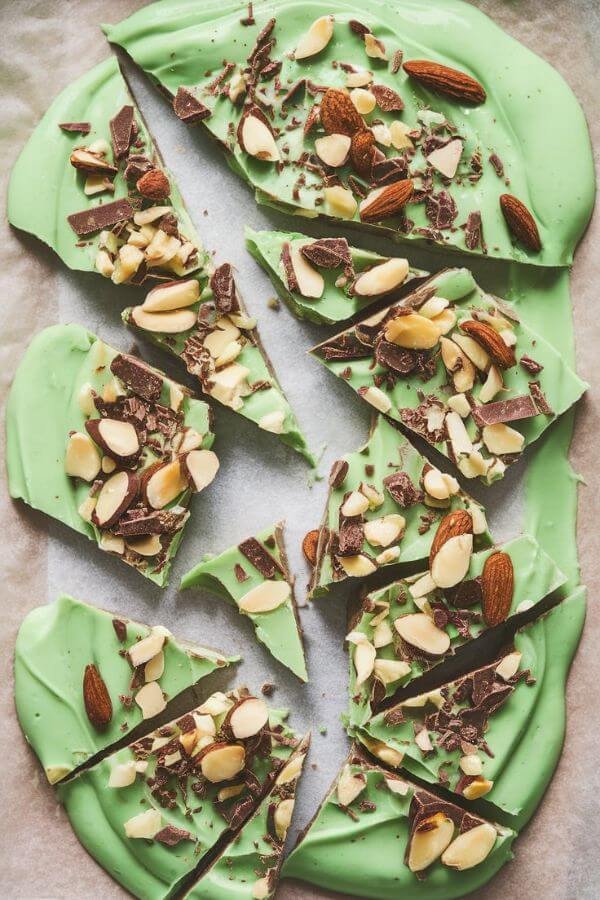 A vibrant green yogurt bark spread thinly on a parchment-lined tray, topped with a scattering of slivered almonds and dark chocolate shavings. The bark is broken into uneven, rustic pieces, with a few displayed on a white board surrounded. The frozen texture and bright colors are highlighted, giving it a fresh and appetizing look.