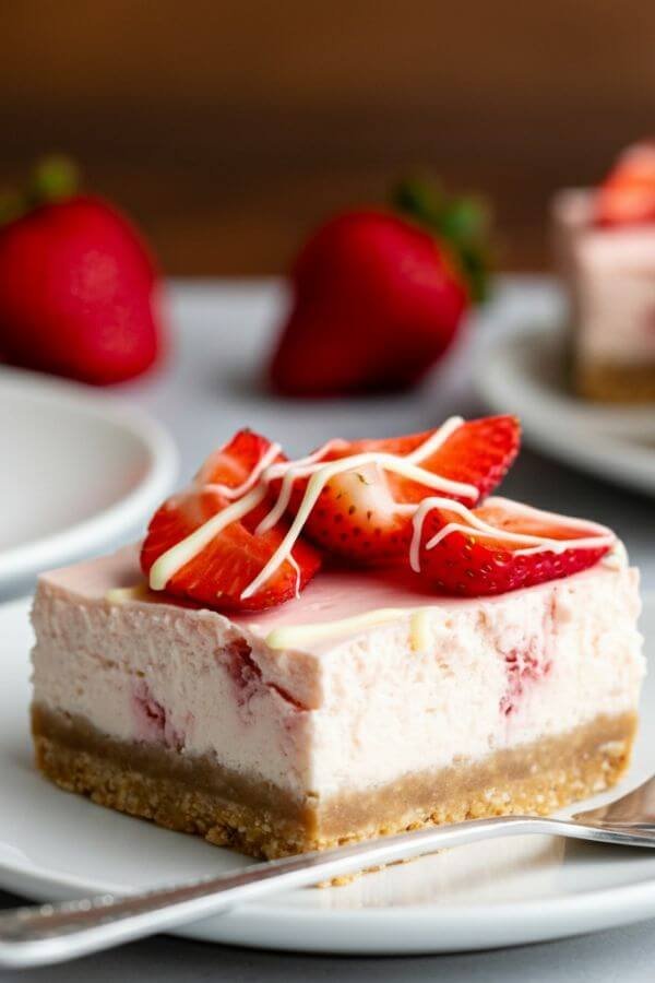 A close-up of strawberry cheesecake bars arranged on a white plate, featuring a golden graham cracker crust, a creamy cheesecake layer, and a vibrant topping of fresh strawberries. Add a drizzle of white chocolate for an elegant touch, with fresh strawberries and a fork in the background for detail.