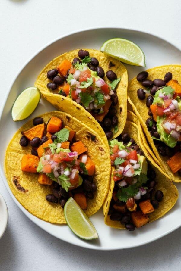 A vibrant plate of tacos filled with roasted sweet potatoes and black beans, garnished with fresh cilantro, lime wedges, and colorful toppings like salsa and avocado. The tacos are arranged on a white table with a small bowl of salsa and a sprinkle of chili powder on the side.