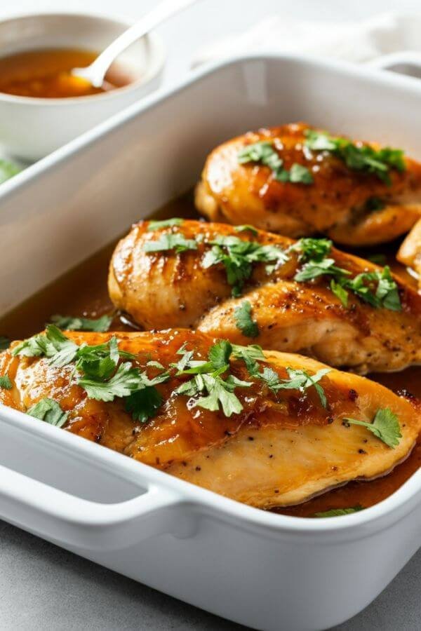 A white baking dish with golden, caramelized chicken breasts, glistening in honey garlic sauce, garnished with fresh parsley or cilantro. Set on a white surface with a small bowl of sauce and a sprinkle of herbs nearby.