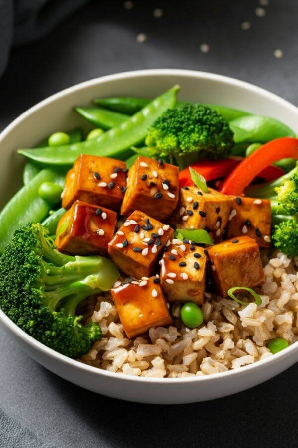 A vibrant bowl featuring golden tofu cubes glazed in teriyaki sauce, surrounded by colorful broccoli, snap peas, and red bell peppers, all atop fluffy brown rice in a white bowl. Perfectly plated for a plant-based meal.