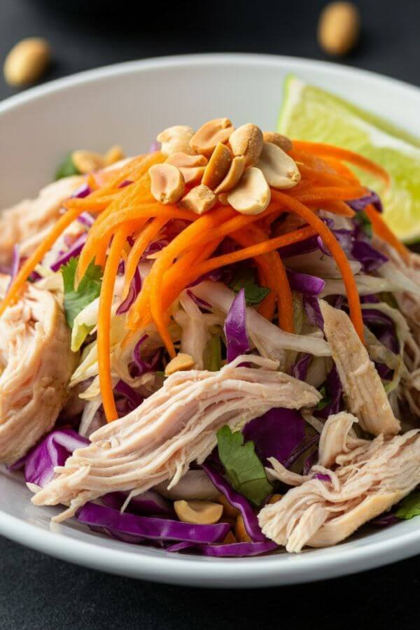 A vibrant shredded Thai chicken salad served in a white bowl, featuring colorful layers of shredded carrots, red cabbage, and chicken. Garnished with chopped peanuts and a lime wedge on the side, the dressing lightly glistens over the fresh ingredients. the chicken is shredded. the salad is mixed