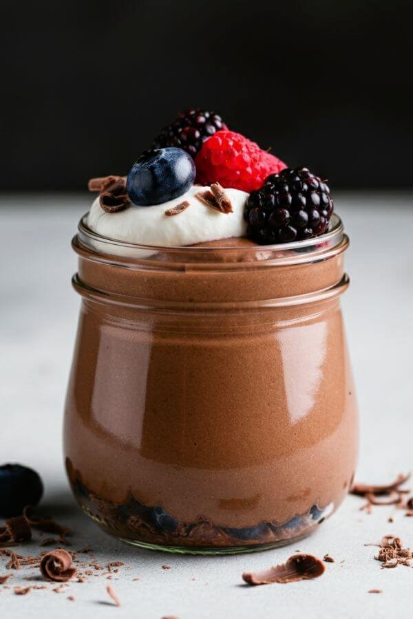 A creamy chocolate mousse served in a glass jar, topped with vibrant fresh berries and a dollop of fluffy coconut whip, placed on a white table with a few scattered chocolate shavings.