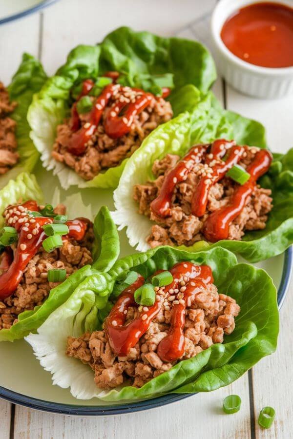 A vibrant plate of crisp green lettuce wraps filled with seasoned ground turkey and topped with a drizzle of sriracha mayo, sprinkled with sesame seeds and chopped green onions, placed on a white wooden table with a small bowl of sauce on the side.