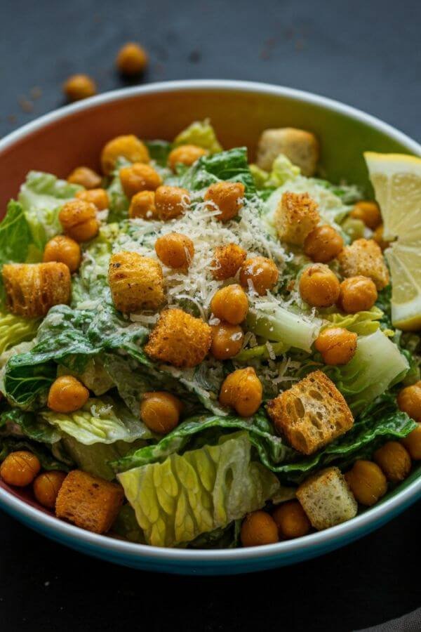 A colorful bowl filled with chickpea pasta, fresh romaine lettuce, crispy roasted chickpeas, golden croutons, and a creamy dressing drizzled over the top. Garnished with a sprinkle of vegan Parmesan and a lemon wedge on the side for a fresh, vibrant look.