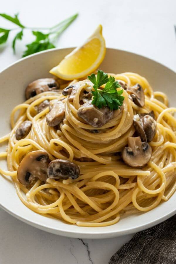 A creamy bowl of linguine pasta coated in a light beige miso mushroom sauce, garnished with fresh parsley and a lemon wedge on the side. The dish is served on a white table with a fork and napkin nearby.