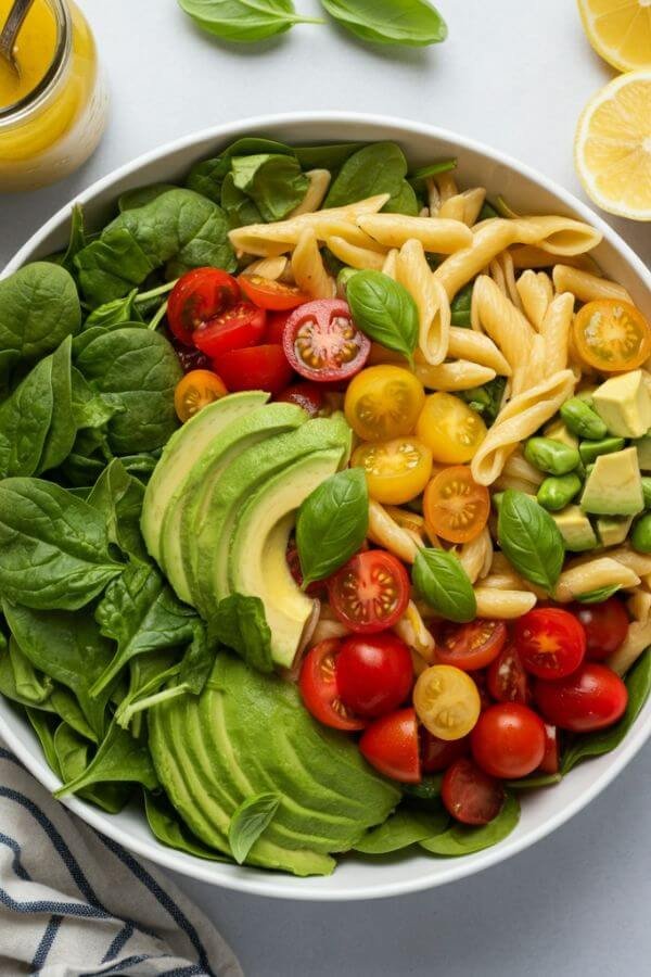 A vibrant overhead shot of a pasta salad in a white bowl, showcasing colorful cherry tomatoes, green spinach, creamy avocado chunks, and basil leaves, with a small jar of lemon garlic dressing and fresh lemons on the side.