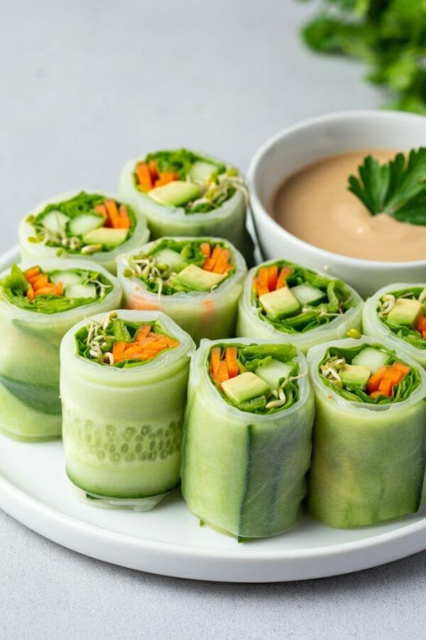 A clean, minimalistic background with a white plate showcasing neatly arranged avocado cucumber rolls. The rolls are tightly wrapped, revealing vibrant layers of green cucumber, creamy avocado, orange shredded carrots, and a hint of sprouts. A small bowl of raw tahini-based dipping sauce sits beside the rolls, garnished with a sprig of parsley. The presentation is fresh, colorful, and inviting, with soft natural light highlighting the textures and details.