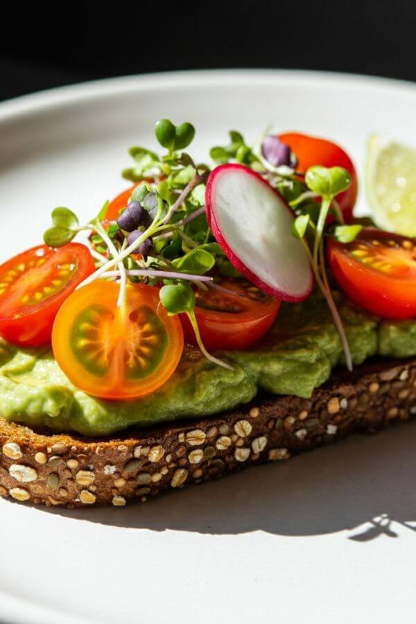 A vibrant slice of whole-grain toast topped with creamy, green avocado spread. Garnished with bright cherry tomato halves, thinly sliced radishes, and fresh microgreens, served on a white dish with a lime wedge on the side.