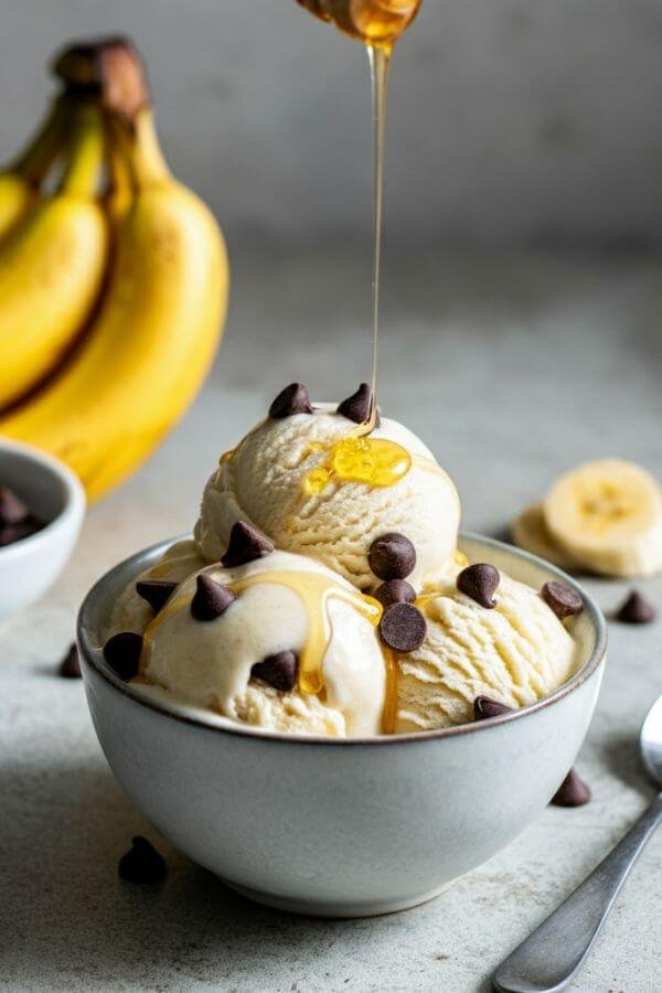 A bowl of creamy banana ice cream topped with chocolate chips and a drizzle of honey, sitting on a white table with a few whole bananas and a spoon nearby.