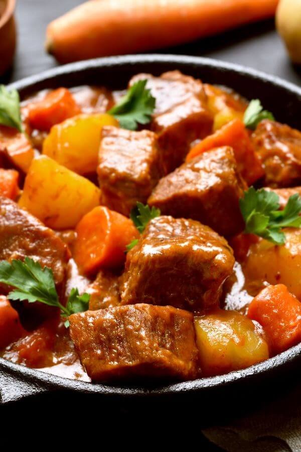 A cozy bowl of beef stew served in a light setting. The dish features tender chunks of beef, vibrant orange carrots, golden potatoes, and a rich, thick gravy. Steam rises from the bowl, hinting at its warmth and comforting aroma. In the background, a loaf of crusty bread sits on a light wooden cutting board, with a knife nearby, ready for slicing. The setting is warm and inviting, with a soft focus on rustic kitchen elements like a wooden table and a linen napkin.