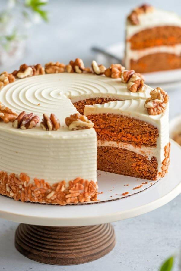 A beautifully frosted carrot cake on a cake stand, with a slice cut to reveal the moist, spiced interior and flecks of grated carrot. Garnish with a sprinkle of chopped walnuts on top for an elegant touch! the background is light and perfect for spring season