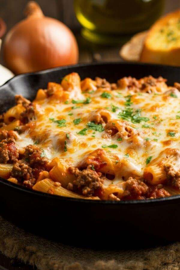 A close-up of a cheesy ground beef pasta skillet. The dish is loaded with golden, melted cheese on top, with tender pasta and savory ground beef mixed in a rich red marinara sauce. The skillet is cast iron, surrounded by a rustic kitchen setting with ingredients like garlic, onion, and fresh parsley scattered around. A side of garlic bread and a sprinkle of shredded cheese complete the cozy, comforting vibe.