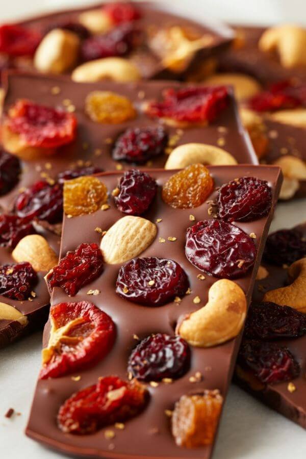 A close-look of glossy, thick smooth chocolate bark broken into irregular pieces, topped with colorful dried fruits, nuts.