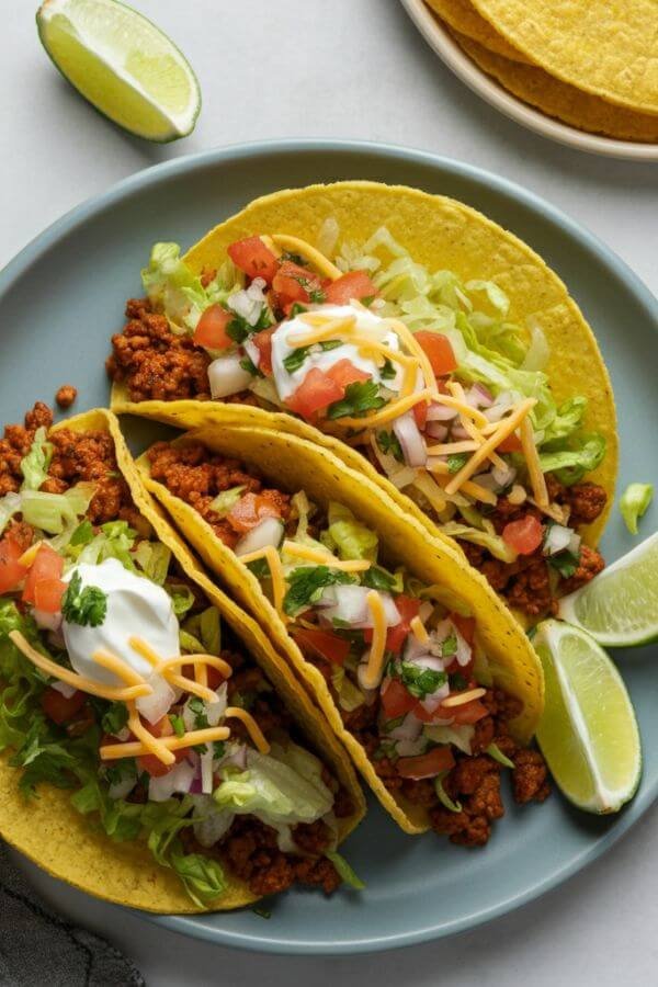 A vibrant plate of tacos filled with juicy seasoned ground beef, topped with crisp lettuce, shredded cheese, fresh diced tomatoes, and a dollop of sour cream, all arranged on a white table with lime wedges and a sprinkle of fresh cilantro.