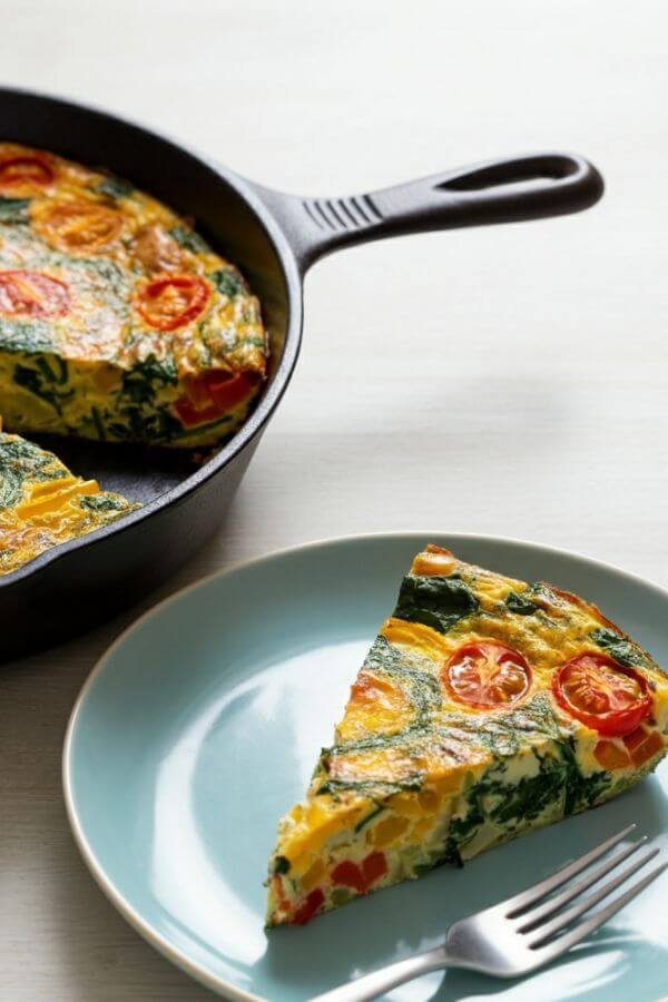 A vibrant and colorful veggie frittata freshly baked in a skillet, golden brown on top with visible chunks of cherry tomatoes, spinach, bell peppers, and melted cheese. A slice is cut and plated nearby, ready to eat! The skillet is placed on a light wooden table. the background is light