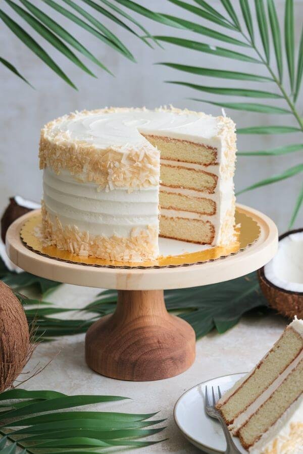 A beautifully frosted coconut cake, topped with a light layer of shredded coconut, placed on a wooden cake stand. The table is adorned with tropical leaves, a few coconuts, and a slice of cake on a white plate to the side, ready to be served. the background is light and perfect for spring season