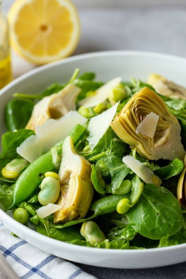 A bright and fresh spring salad in a white bowl, featuring vibrant green fava beans, artichokes, and mixed greens. Shaved Parmesan slices are sprinkled on top, with a drizzle of lemon vinaigrette glistening on the veggies. The setting is light and airy, with a slice of lemon and a small jar of vinaigrette in the background for a fresh, homemade touch.