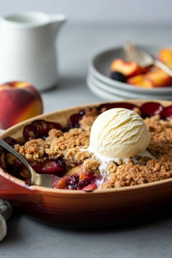 A warm, golden-brown fruit crumble fresh out of the oven, served in a baking dish with a scoop of vanilla ice cream melting on top. Include vibrant fruits like apples, peaches, or berries peeking through the crispy topping, with a rustic, cozy kitchen setting in the background.