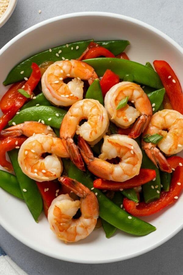 A vibrant overhead shot of a Garlic Shrimp Stir-Fry served in a white bowl. Plump, pink shrimp are nestled among colorful bell peppers and snap peas, glistening with soy sauce. The dish is garnished with a sprinkle of sesame seeds and fresh herbs.