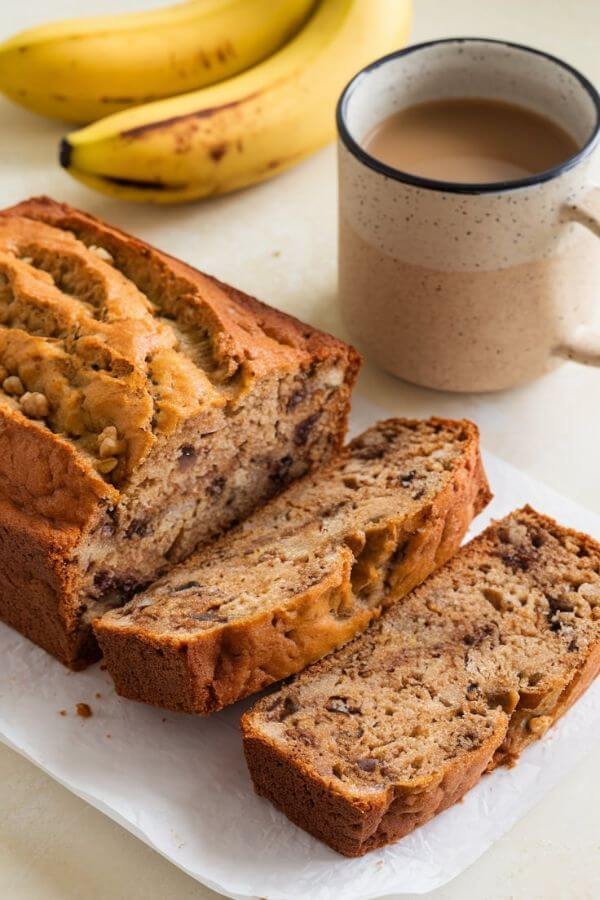 A loaf of golden-brown banana bread on a white table. Slices reveal its moist texture, with visible bits of walnuts or chocolate chips, alongside a mug of coffee and a few ripe bananas in the background.