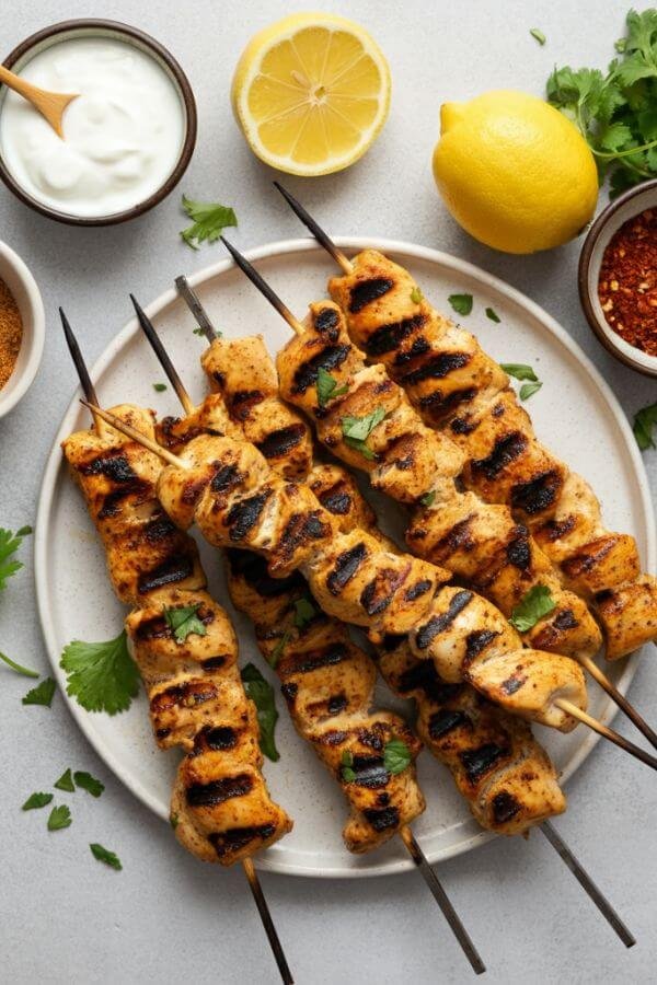 A vibrant, overhead shot of Greek yogurt marinated chicken skewers on a grill, with golden-brown char marks, surrounded by small bowls of ingredients like Greek yogurt, paprika, garlic powder, and lemon slices. The skewers are placed on a white dish next to fresh herbs like parsley or cilantro for garnish, with a light, natural background that highlights the freshness and simplicity of the dish.