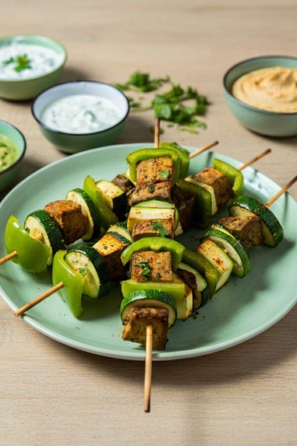 A vibrant plate of skewers with green vegetables carefully arranged in a pattern, served with small dipping bowls of tzatziki, ranch, and hummus. Set on a light wooden table with fresh herbs scattered around for extra color.