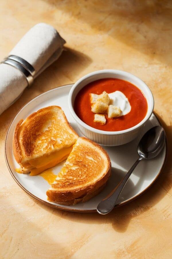 Create a cozy, inviting close look of flat-lay image of a grilled cheese and tomato soup setup. The grilled cheese should be golden-brown with melted cheddar oozing slightly from the edges, cut diagonally to show the cheesy interior. Place the sandwich on a white plate next to a bowl of rich, vibrant tomato soup garnished with a dollop of sour cream and a few crunchy croutons. Add a spoon next to the bowl and a folded napkin for a homely touch. The background should be a warm, textured surface, like a lightnwooden table, with soft natural lighting to emphasize the warmth and comfort of the meal.