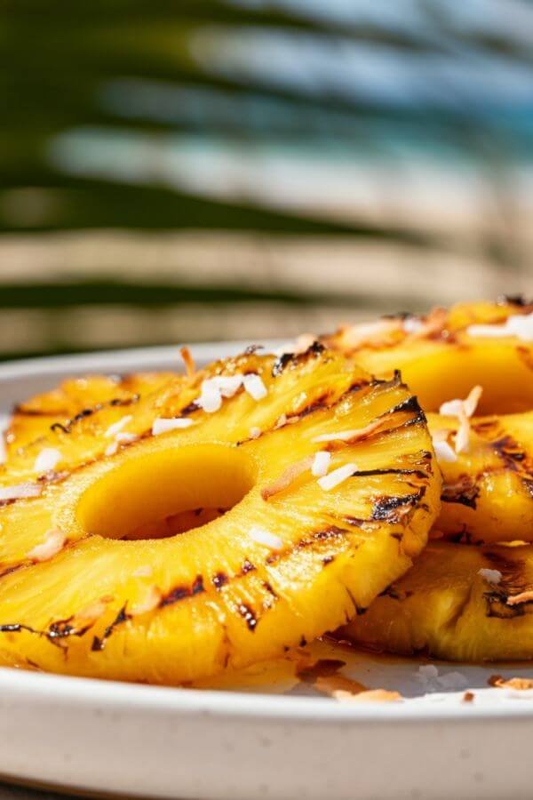 A close-up of caramelized pineapple slices on a grill, with golden grill marks. The slices are plated on a white dish and topped with toasted shredded coconut, set against a bright, summery background with hints of palm leaves or a beachy vibe.