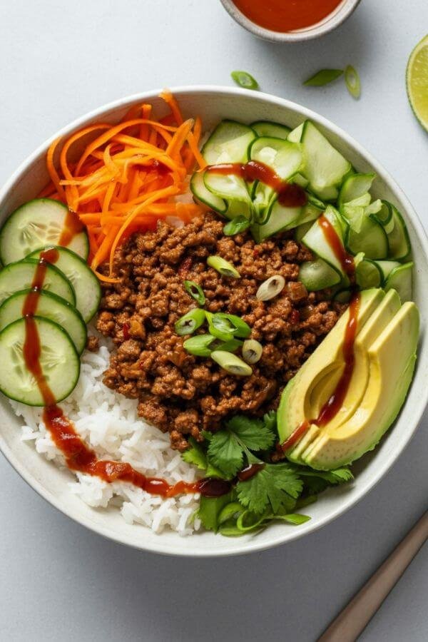 A vibrant, top-down view of a Ground Beef and Rice Bowl. The bowl features a base of fluffy white rice topped with perfectly browned ground beef glistening with soy sauce and lime juice. Fresh, colorful toppings include sliced cucumber, shredded carrot, and creamy avocado slices arranged neatly around the bowl. A drizzle of sriracha or creamy dressing adds a finishing touch. Bright, natural lighting highlights the textures and colors, with a clean, minimal background to keep the focus on the dish. Include a spoon or chopsticks resting beside the bowl for a casual dining feel.