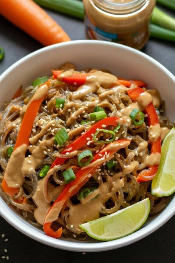 A vibrant, healthy dish of Kelp Noodle Pad Thai served in a large bowl. The kelp noodles glisten under the light, mixed with colorful julienned carrots, thinly sliced red bell peppers, and chopped green onions. A creamy almond butter sauce lightly coats the noodles, adding a rich texture. Garnish with a sprinkle of sesame seeds and a lime wedge on the side for a fresh, appetizing look. The plate is styled on a white dish with natural lighting, surrounded by the raw ingredients—kelp noodles, a whole carrot, red bell pepper, green onions, almond butter, and lime.