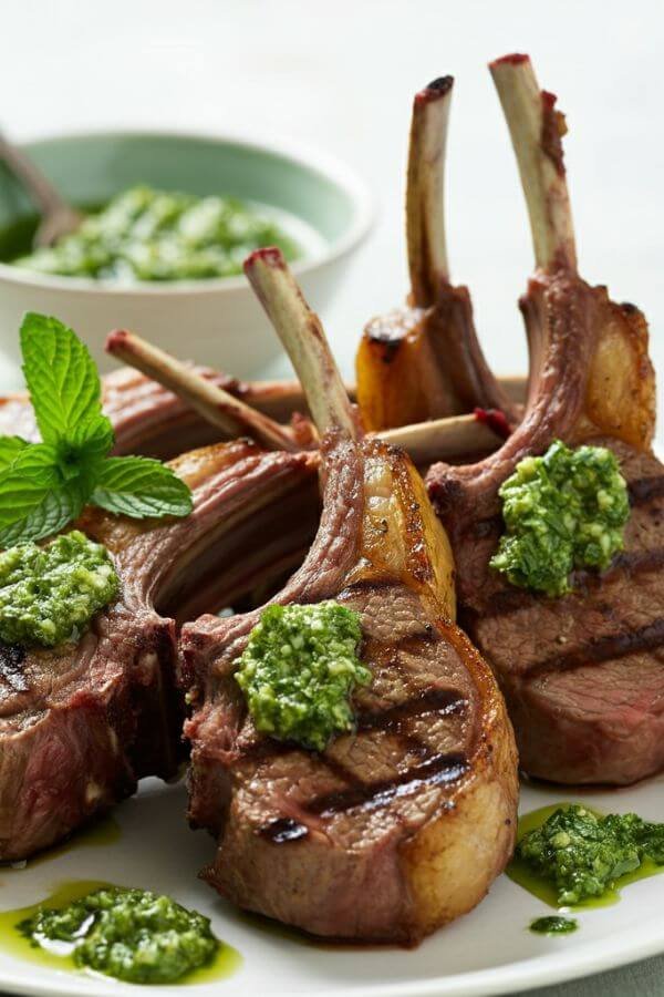 A close look of a perfectly grilled lamb chops drizzled with vibrant green mint pesto, garnished with a sprig of fresh mint. On the side, a small bowl of pesto and a glass of red wine complete the inviting springtime setting.