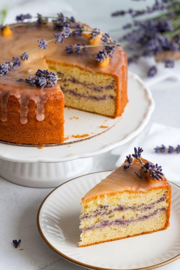 A beautifully styled Lavender Honey Cake placed on a white dish. The cake is golden brown with a light glaze, showcasing its soft texture and delicate crumb. A slice of the cake is cut and placed on a white plate nearby, revealing its moist interior. the background is light and perfect for spring season.