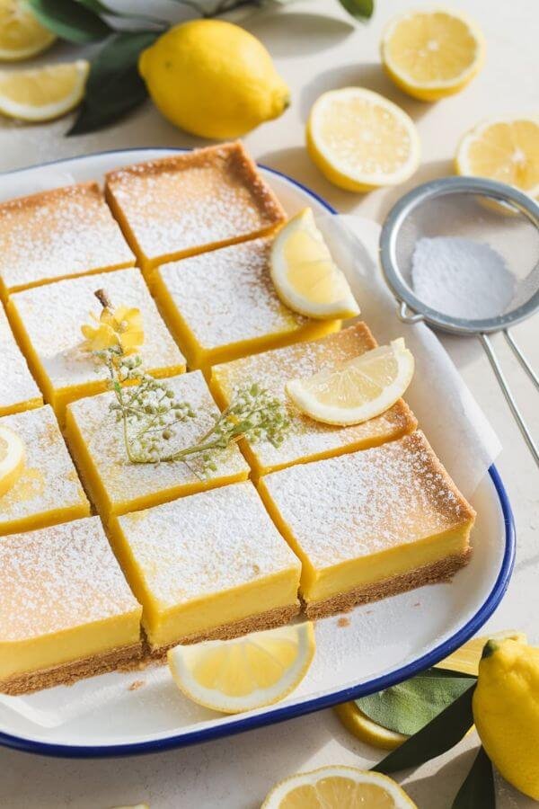 A close top-down view of a tray of freshly baked lemon bars on a white dish. The bars are cut into neat squares, with a golden crust and a vibrant yellow filling. A light dusting of powdered sugar covers the tops, giving a soft, snowy appearance. Surround the tray with a few scattered lemons, lemon slices, and sprigs of greenery for a fresh and natural look. Add a small sieve with powdered sugar beside the tray to hint at the finishing touch. The lighting should be soft and natural, emphasizing the bright, sunny tones of the dish.