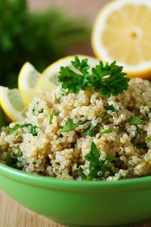 A close look of A vibrant bowl of fluffy quinoa mixed with fresh green parsley, glistening with olive oil, and garnished with thin lemon slices on the side. The dish is displayed on a light wooden table with fresh herbs and a cut lemon in the background.