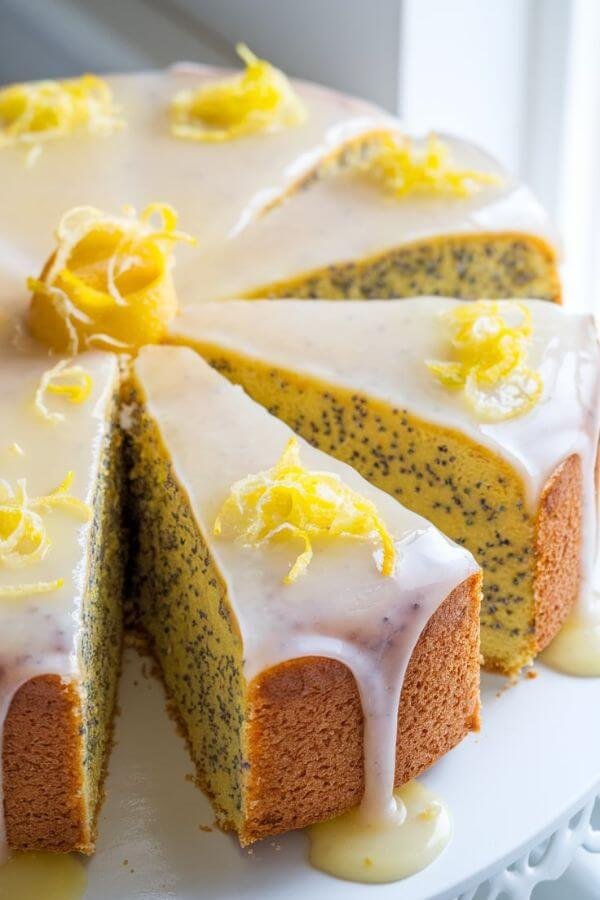 A freshly baked Lemon Poppy Seed Cake placed on a white dish, cut into slices to reveal its moist and buttery texture with visible poppy seeds. The cake is topped with a glossy lemon glaze that drips slightly down the sides, garnished with a sprinkle of fresh lemon zest. Bright, natural lighting enhances the zesty and fresh springtime feel.