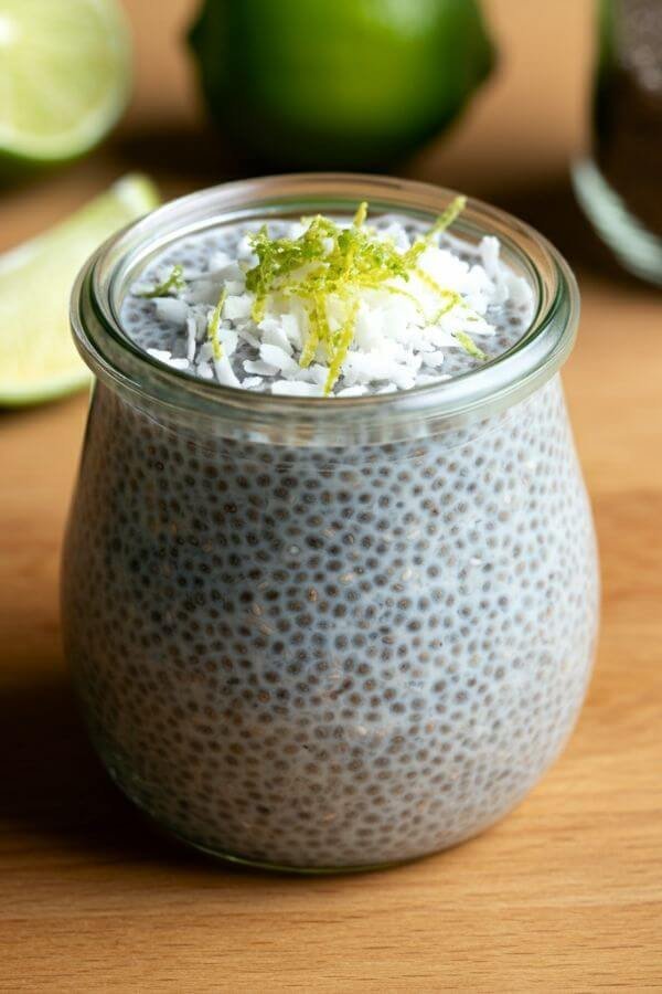 A glass jar filled with creamy pudding, topped with a sprinkle of shredded coconut and lime zest, placed on a light wooden table with fresh lime slices and a small jar of chia seeds in the background.