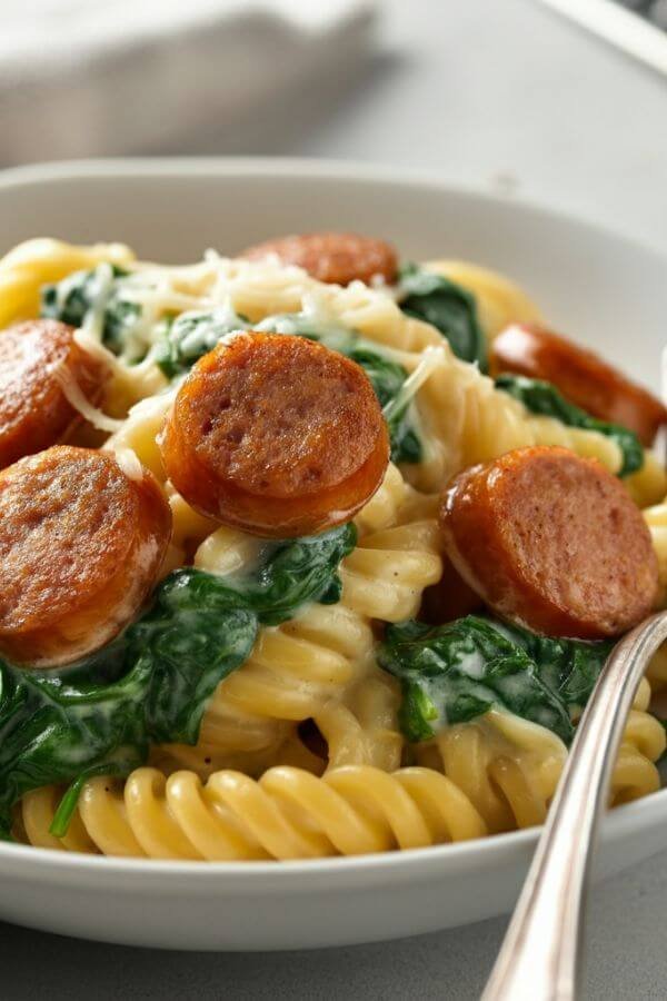 A bowl of creamy pasta featuring golden-brown rounds of maple sausage, bright green spinach, and a rich, cheesy sauce. Garnished with extra Parmesan and served with a fork on a white table.