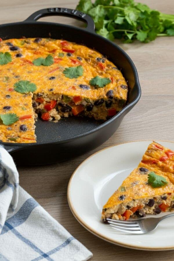 A close look of A vibrant and appetizing Mexican-inspired frittata freshly baked in a skillet, with a golden, cheesy top. The frittata is packed with colorful ingredients like diced red and green bell peppers, black beans, and fresh cilantro sprinkled on top. A slice is cut and plated nearby, ready to eat!. It’s served on a light wooden table. The warm, inviting lighting highlights the textures and bold colors, making it feel both homely and delicious.