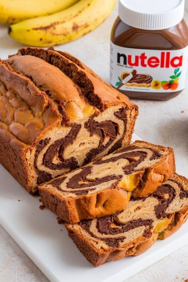 A loaf of Nutella Banana Bread sliced to reveal the beautiful Nutella swirls inside. The bread is golden brown on the outside and moist on the inside, served on a white cutting board with a small jar of Nutella and ripe bananas in the background.