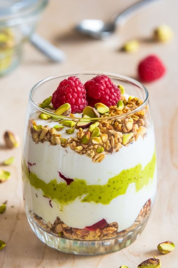 A clear glass filled with visible layers of creamy yogurt, vibrant green pistachio cream, and golden granola. The top is garnished with chopped pistachios, a few fresh raspberries, and a light drizzle of honey. The background features a light wooden table with a spoon and some scattered pistachios.