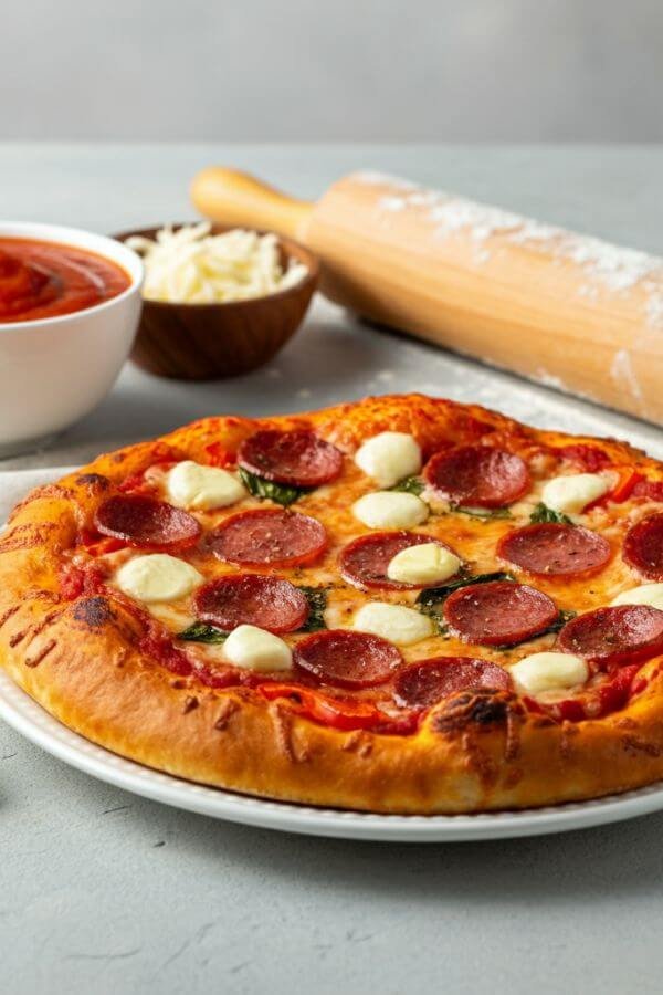 A freshly baked homemade pizza on a white dish. The pizza has a golden, crispy crust brushed with garlic butter, bubbling mozzarella cheese, and a variety of colorful toppings like pepperoni and vegetables. Next to the pizza, there are ingredients like a bowl of marinara sauce, shredded mozzarella, fresh basil, and a rolling pin with some scattered flour, emphasizing the homemade preparation.