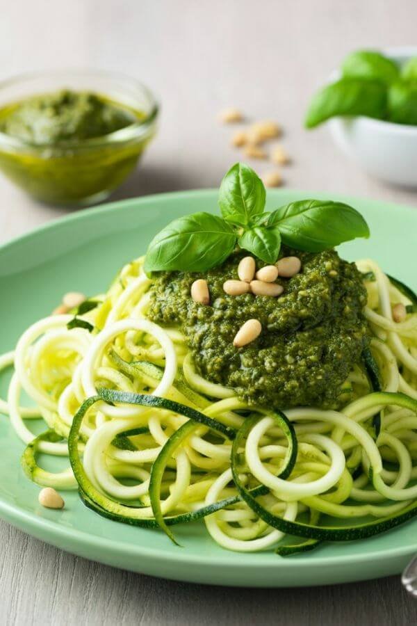 A vibrant plate of raw zucchini noodles ("zoodles") tossed in a creamy, green basil pesto sauce, topped with fresh basil leaves and a sprinkle of pine nuts. The dish is set on a light wooden table with a small bowl of pesto, a handful of fresh basil, and scattered pine nuts in the background for a fresh, wholesome look. The lighting is natural and soft, emphasizing the bright green hues and fresh ingredients.