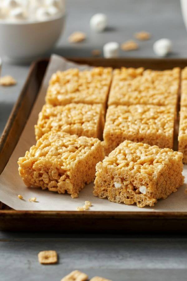 A tray of golden Rice Krispie Treats sitting on a white countertop. The treats are cut into neat squares, with a few scattered mini marshmallows and a bowl of Rice Krispies cereal placed nearby for a cozy, nostalgic vibe.