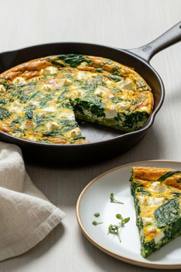 A close look of A golden-brown frittata with visible chunks of creamy feta and vibrant green spinach, served in a skillet on a light wooden table, garnished with a sprig of fresh herbs. A slice is cut and plated nearby, ready to eat! the background is light